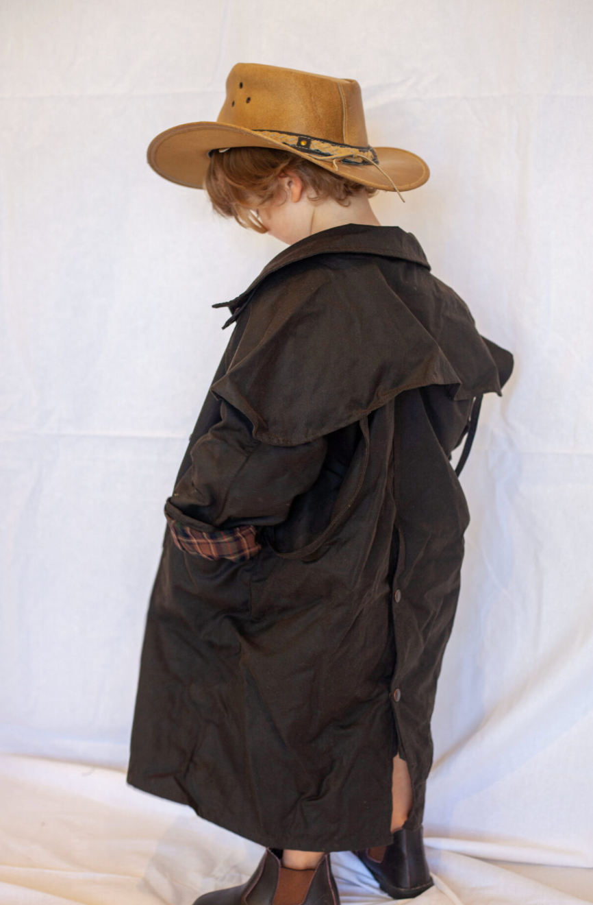 A person wearing a dark brown waxed canvas trench coat, a tan cowboy hat, and brown boots, photographed from the back against a white fabric backdrop.