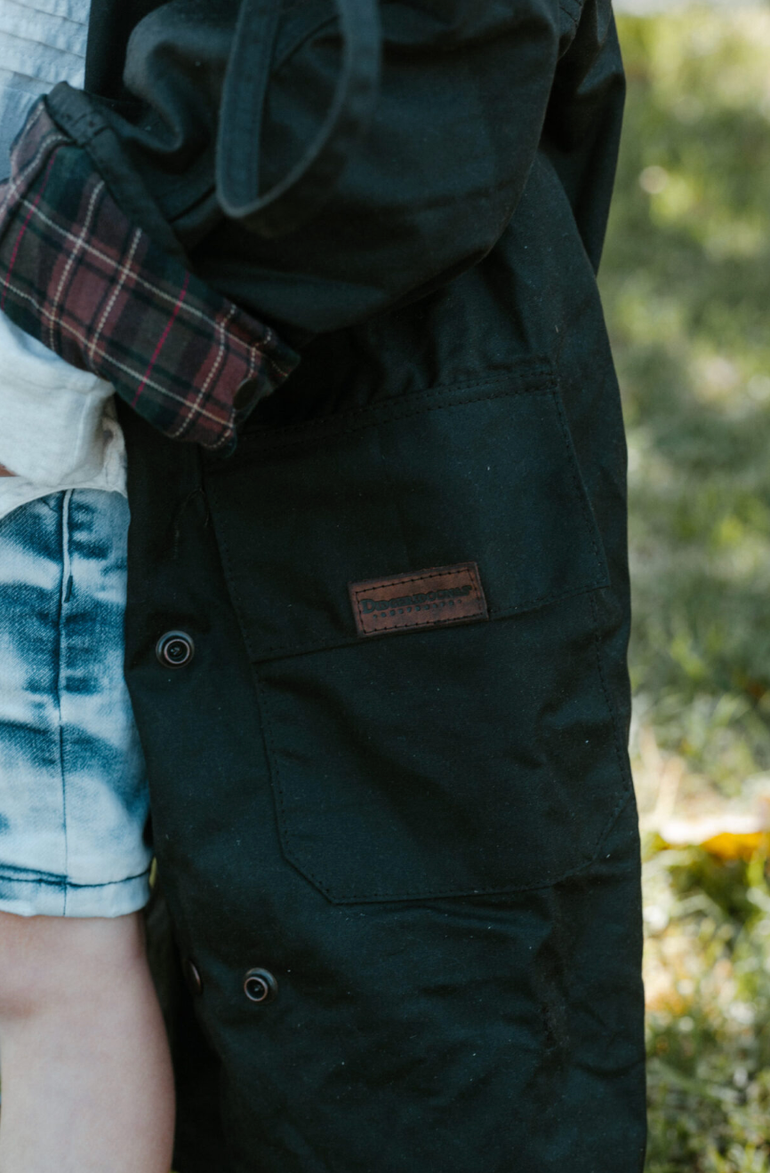 A close-up outdoor shot of a person wearing a dark green utility jacket with a brown leather patch on the pocket, paired with a plaid shirt and denim shorts.