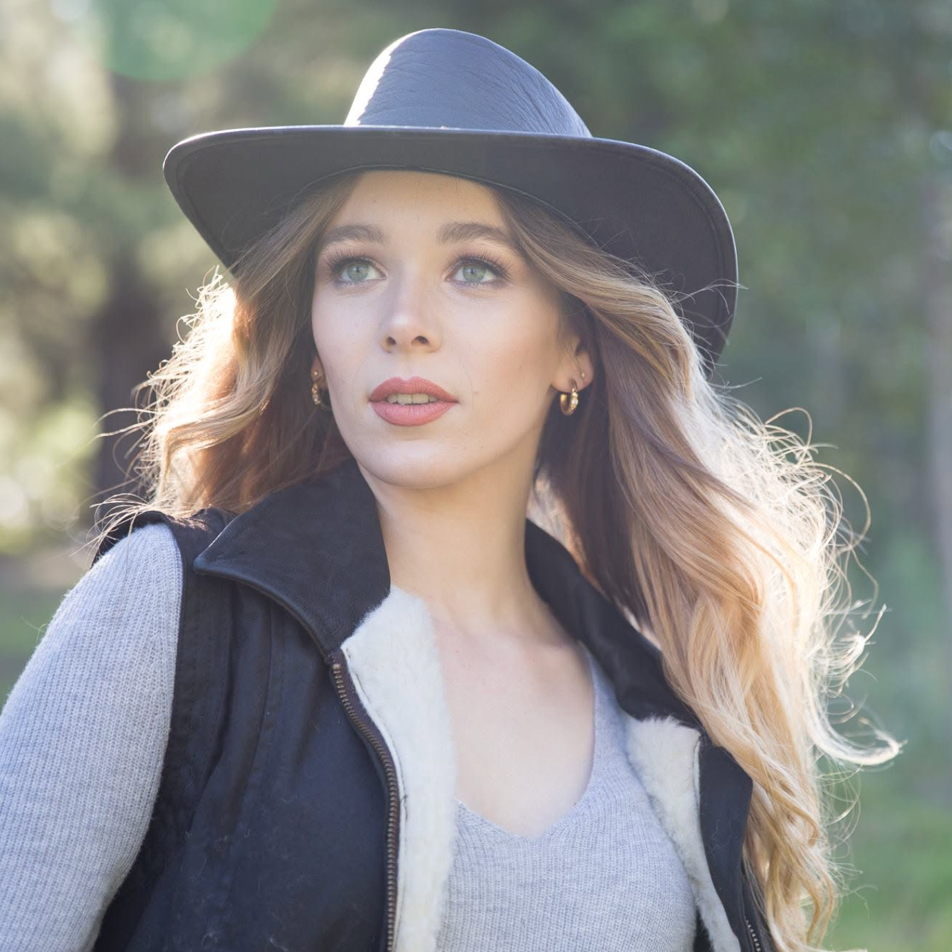 A woman outdoors wearing a wide-brim black hat, a black vest with white sherpa lining, and a gray knit top. The background is a softly blurred natural green setting.