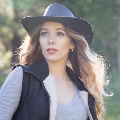 A woman outdoors wearing a wide-brim black hat, a black vest with white sherpa lining, and a gray knit top. The background is a softly blurred natural green setting.