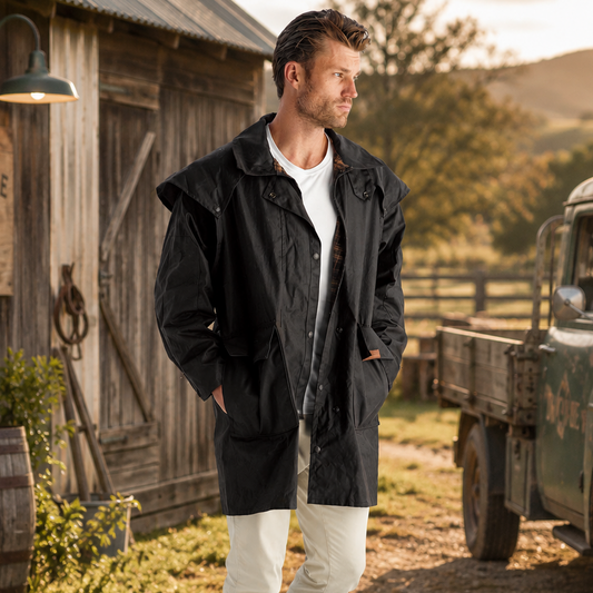 A man stands outdoors in front of a rustic wooden barn and vintage truck, wearing a black hooded jacket over a white shirt with off-white pants and black boots. A sign on the barn reads 'Countryside Gear'.
