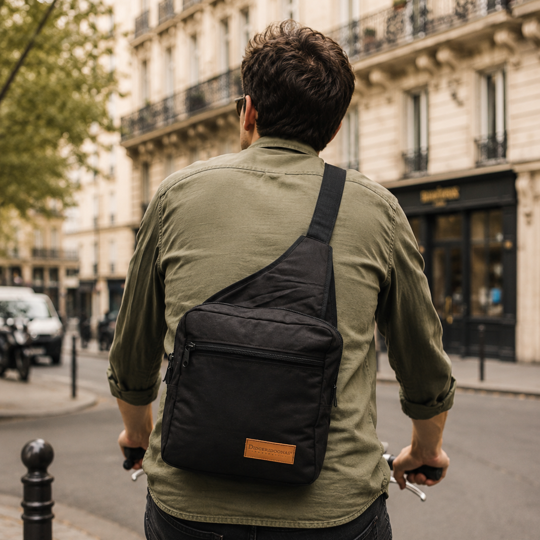 A man riding a bicycle in a city street, viewed from behind, wearing a black diagonal crossbody sling bag with a small tan leather patch on the front pocket.