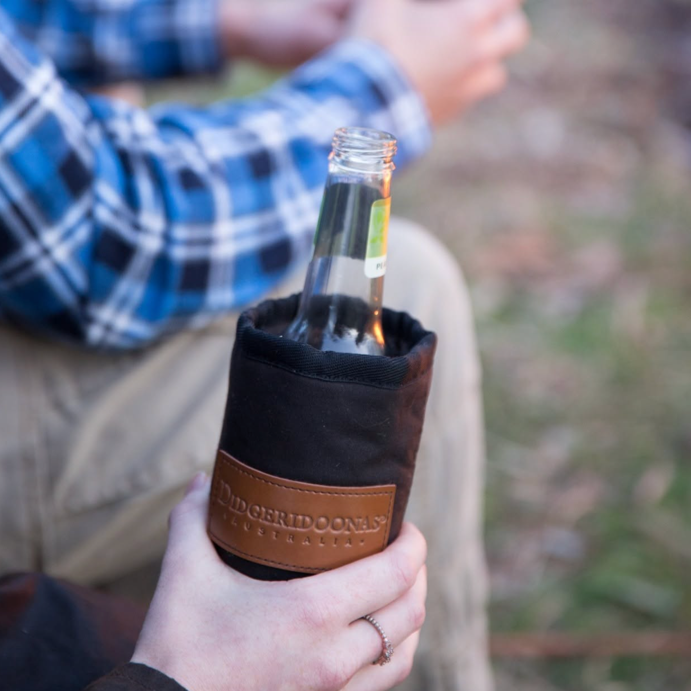A person outdoors holding a clear glass bottle inside a black insulated bottle sleeve with a brown leather band. The sleeve has a visible embossed brand patch reading 'The Orchid Company'.