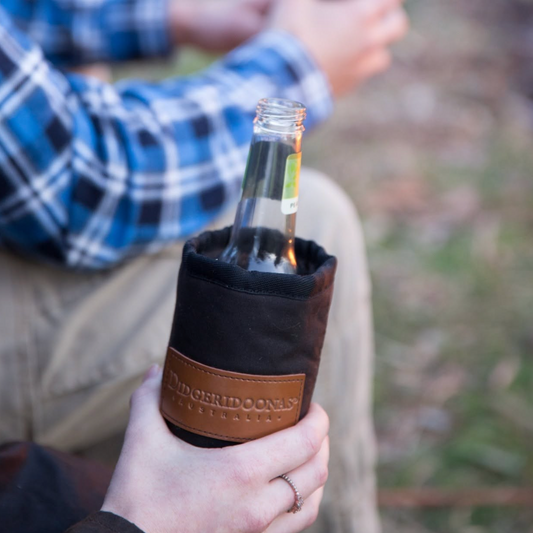A person outdoors holding a clear glass bottle inside a black insulated bottle sleeve with a brown leather band. The sleeve has a visible embossed brand patch reading 'The Orchid Company'.