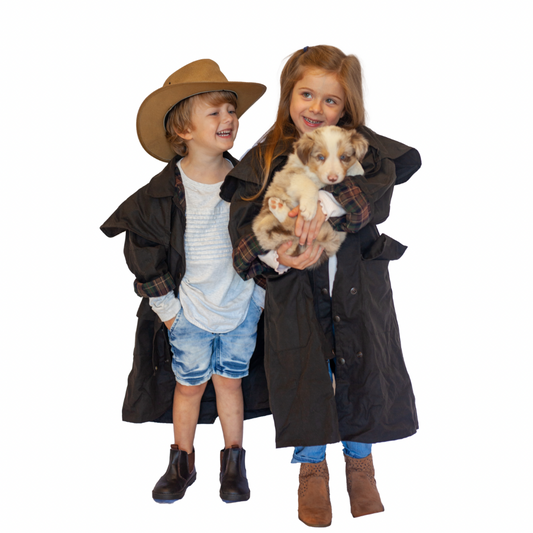 Two children wearing full-length black coats, standing and smiling while holding a small dog.