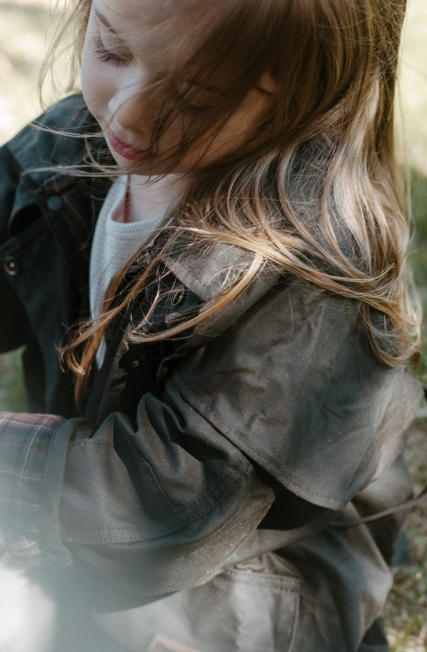 A close-up lifestyle shot of a young girl with long light brown hair wearing a dark, weathered brownish-gray jacket over a white shirt, captured in soft, natural light.